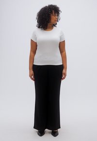 Λευκή μπλούζα με κοντά μανίκια συνδυασμένη με μαύρο παντελόνι φαρδιάς γραμμής. Το μοντέλο στέκεται όρθιο, παρουσιάζοντας την εφαρμογή και τη ροή της στολής. Απλός σχεδιασμός και χρώματα.