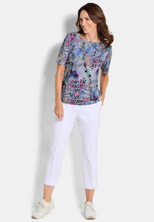 Woman standing with hands in pockets, wearing a colorful patterned short-sleeve top, white cropped pants, and white sneakers.