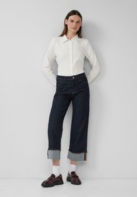 Witte blouse met lange mouwen en knopen, gecombineerd met donkerblauwe denim brede broek, met een omgeslagen zoom en subtiele blauwe en gele stikselaccenten.
