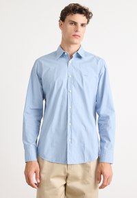 Camisa de manga longa azul claro, feita de algodão, com colarinho abotoado, um pequeno logotipo bordado e botões brancos na parte da frente e nos punhos.