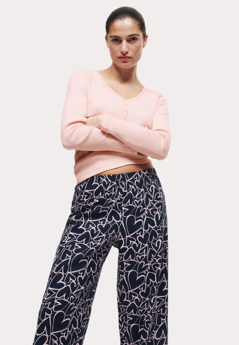Helles rosa Langarmshirt mit V-Ausschnitt und Knöpfen, kombiniert mit dunkelblauen Hosen mit einem weißen Herzmuster. Weicher Stoff, lässige Passform.