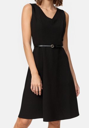 Ärmelloses schwarzes knielanges Kleid mit V-Ausschnitt und schmalem schwarzen Gürtel mit runder Schnalle an der Taille, getragen von einer stehenden Person.