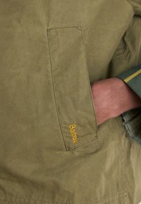 Casaco verde oliva com um bolso visível e o logotipo da Barbour em bordado laranja, feito de um tecido rugoso e texturizado.