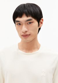 Helles cremefarbenes Sweatshirt aus weichem Stoff mit rundem Halsausschnitt und einem dezenten gestickten Logo auf der linken Brust.