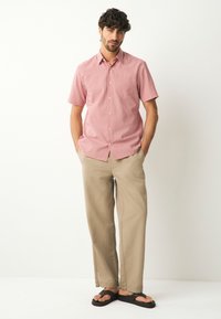 Chemise rouge à manches courtes avec des pois blancs, associée à un pantalon beige ample et des tongs noires, posant devant un fond neutre.