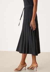 Donna vestita con una gonna midi plissettata nera con vita legata, abbinata a sandali con tacco alto sabbia su uno sfondo neutro.