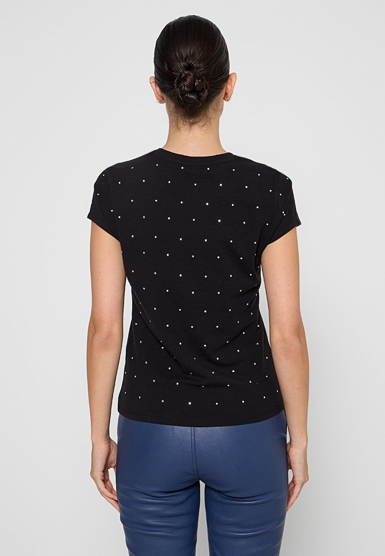 Femme avec un chignon portant un tee-shirt noir à manches courtes avec de petits points réfléchissants et un pantalon en cuir bleu, vue de dos.