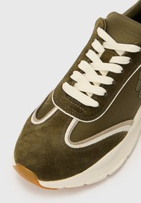 Zielone sneakersy w kolorze oliwkowym z zamszową cholewką, białymi sznurówkami i kontrastowymi beżowymi akcentami. Gruba podeszwa z teksturowaną gumową podeszwą.