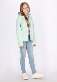 Giacca verde menta chiaro con cappuccio, frontale con zip e fodera interna a contrasto, abbinata a jeans azzurri e sneakers bianche.
