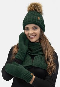 Cappello lavorato a maglia verde con un pompon in pelliccia sintetica, sciarpa abbinata con un motivo a trama, e muffole. Include un piccolo dettaglio del logo in pelle.