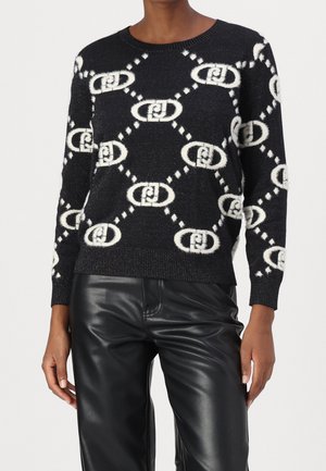 Persona con un suéter negro con patrón de cadenas ovaladas blancas y pantalones de cuero negros, de pie contra un fondo claro y liso.