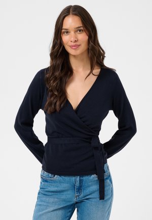 Woman with long wavy brown hair wears a navy wrap sweater and blue jeans, standing against a plain light background, hands behind her back.