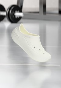 Baltas slip-on apavi ar gludu tekstūru, ergonomisku formu un ventilācijas atverēm, ar smalku iegravētu logotipu sānos.