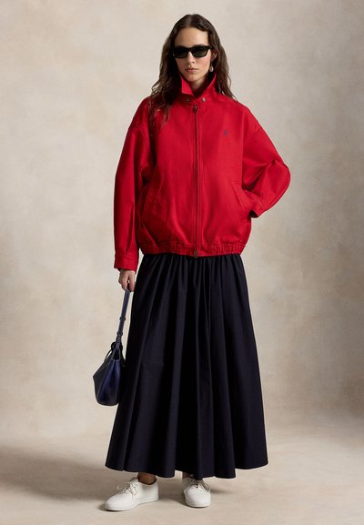 Femme portant des lunettes de soleil noires, une veste zippée rouge, une longue jupe noire, des baskets blanches, et tenant un petit sac à main foncé sur un fond neutre.