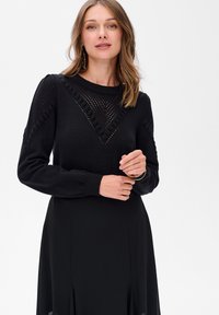 Czarny sweter o dzianinowej fakturze, z wzorzystą łatą i prążkowanymi mankietami. Modelka nosi go z czarną spódnicą, prezentując luźny krój.