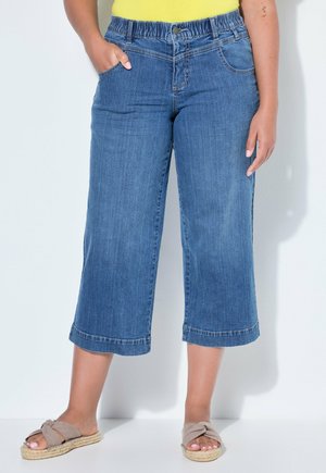 Person iført korte blå denimjeans med elastisk taljebånd og beige slip-on sandaler, stående mod en ensartet lys baggrund.