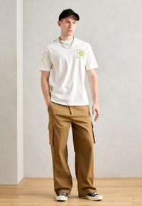 White cotton T-shirt with green graphic on left chest, paired with tan cargo pants featuring side pockets. Black cap and sneakers complete the look.