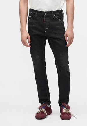 Mand iført sorte jeans med lige ben, hvid t-shirt og mørkerøde sneakers med beige snørebånd, står mod en ensfarvet baggrund.