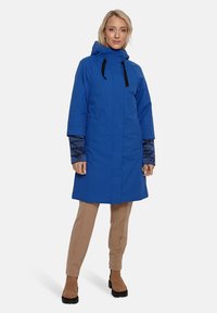 Zils ūdensizturīgs mētelis ar kapuci, garām piedurknēm un melniem akcentiem. Iezīmējas ar gludu tekstūru un brīvu piegulumu. Valkāts virs beigu biksēm un zābakiem.