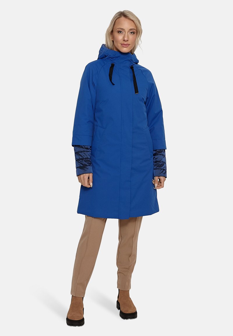 Zils ūdensizturīgs mētelis ar kapuci, garām piedurknēm un melniem akcentiem. Iezīmējas ar gludu tekstūru un brīvu piegulumu. Valkāts virs beigu biksēm un zābakiem.