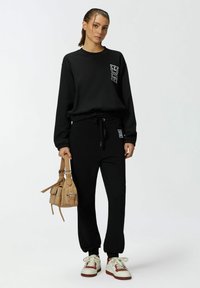 Zwarte sweatshirt met logo-detail, bijpassende zwarte joggers en witte sneakers. Het model houdt een kleine beige handtas vast.