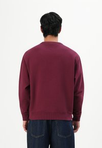 Hombre de pie de espaldas, vistiendo un sudadera burgundy lisa y jeans oscuros contra un fondo blanco.