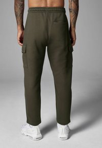 Olivenfarbene Cargo-Hosen mit einem Kordelzugbund, seitlichen Taschen und einer Gesäßtasche, kombiniert mit weißen Sportschuhen. Glatter Stofftextur.