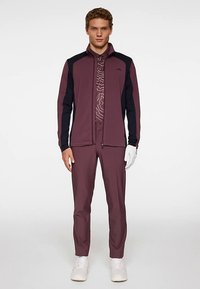 Vinröd jacka med svarta detaljer, dragkedja framtill och hög krage, kombinerad med vinröda byxor. Innehåller en mönstrad skjorta under och vita sneakers.