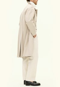Trench coat beige con colletto alto, tessuto leggero, tasche laterali, abbinato a pantaloni crema e mocassini neri lucidi.