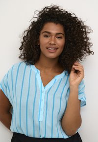 Chemise bleu clair à manches courtes avec des rayures verticales blanches, décolleté en V et fermeture à boutons. Fabriquée en tissu léger, avec une texture douce.