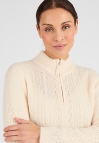 Suéter de cremallera de color beige claro, elaborado con material suave, que presenta un patrón de punto texturizado con detalles de trenzado y calados en las mangas.
