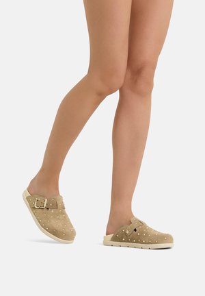 Gammbre con zoccoli beige slip-on con fibbia e dettagli con borchie, in piedi su sfondo bianco.