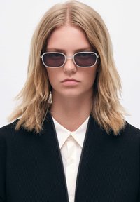 Suot rectangular dengan lensa abu-abu dan bingkai logam, dipakai oleh seseorang dengan rambut pirang, dipadukan dengan jaket hitam bergaris dan kemeja putih.