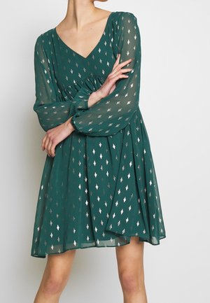 Woman wearing a knee-length green dress with silver diamond patterns, long sheer sleeves, and a V-neck, posing with arms crossed.