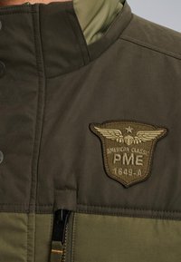 Olivgrön jacka med ett lagerdesign, som har en patch med vingar, dragkedjestängning och textilmaterial. Accenter i en mörkare grön nyans.