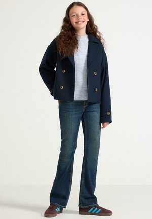 Marineblauwe dubbelzijdige peacoat met gouden knopen, gedragen over een lichtgrijze trui. Gecombineerd met flare donkerblauwe jeans en bruine sneakers met blauwe accenten.