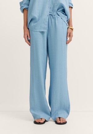 Frau trägt lockere hellblaue Leinenhose und ein passendes Hemd, steht barfuß vor einem schlichten hellen Hintergrund.