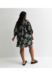 Schwarzes Kleid mit weißem Blättermuster, mit puffärmeln und lässigem Schnitt, das bis zur Mitte des Oberschenkels reicht. Wird mit schwarzen Sandalen getragen.