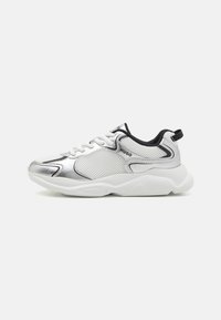 Zapatillas de malla blancas con acentos plateados, ribetes negros y cordones blancos. El diseño presenta una forma redondeada con una suela texturizada y detalles del logotipo.