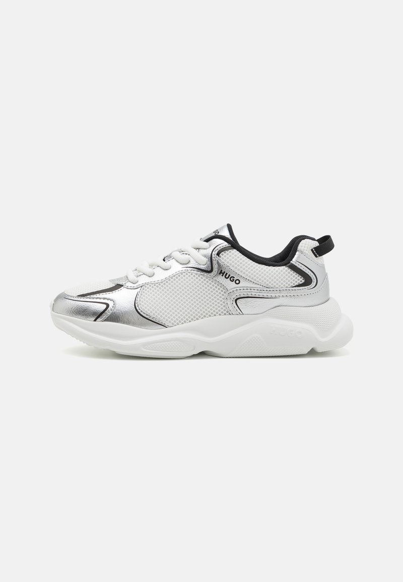 Zapatillas de malla blancas con acentos plateados, ribetes negros y cordones blancos. El diseño presenta una forma redondeada con una suela texturizada y detalles del logotipo.