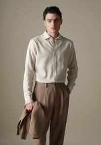Hellbeige Leinenhemd mit Button-Down-Kragen, kombiniert mit braunen, strukturierten Hosen. Hält einen braunen Pullover mit gerippten Bündchen.