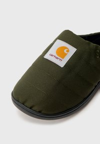 Zielony pikowany but wsuwany z teksturowaną tkaniną, czarną podeszwą oraz białą naszywką z logo ozdobioną pomarańczowymi i żółtymi akcentami.