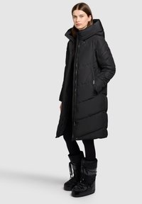 Svart vadderad jacka med hög krage och stor huva. Slät yta, lång modell med sidofickor. Kombinerad med svarta snöskor.