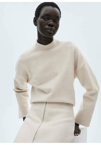Hellbeiger Pullover mit hohem Kragen, lässiger Passform, gerippten Bündchen und nahtlosem Design. Kombiniert mit einem fließenden beigen Rock mit einem seitlichen Schlitz.