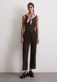 Braune Wildleder-Hosen in hoher Taille mit Vorder- sowie Taschen, kombiniert mit einem dunklen Tanktop und einem hellpinkfarbenen gemusterten Schal. Schwarze Slipper runden das Outfit ab.
