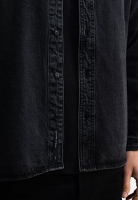 Zwarte denimshirt met knoopsluitingen, een gestructureerde stof en een binnenlabel. Draagt over een zwart shirt, wat gelaagde stijlen toont.