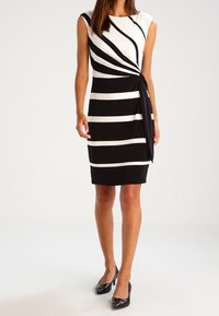 Abito a righe bianche e nere con maniche corte, un nodo drappeggiato di lato e una silhouette aderente. Il tessuto è liscio con una leggera lucentezza.