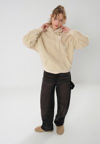 Felpa beige con cappuccio e tasca frontale, abbinata a pantaloni larghi scuri e stivali color cuoio. Vestibilità rilassata con texture.