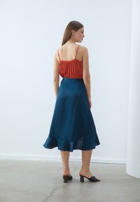 Rød plissert cami kombinert med et flytende teal satin skjørt med bølgete kant. Modellen har på seg svarte glidelysandaler med minimalistisk design.