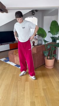 Rode trainingsbroek met witte zijkanten, gecombineerd met een lichtgrijs t-shirt en zilveren sportschoenen. Binnenomgeving met planten en meubels.